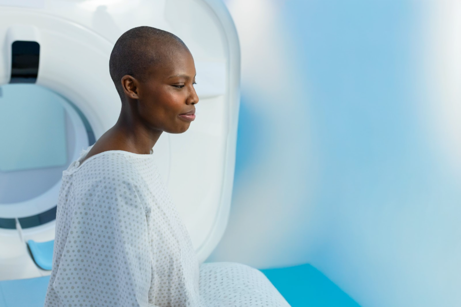 Woman receiving cancer treatment sits beside a medical imaging machine, wearing a hospital gown.